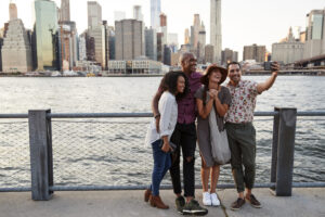 Vier Personen machen am Wasser vor der Skyline von New York ein Selfie mit dem Smartphone