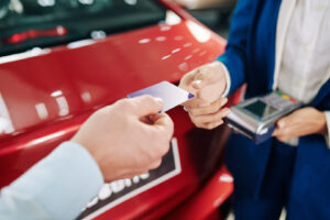Kunde übergibt einem Mitarbeiter mit Kartenlesegerät seine Kreditkarte neben einem Auto.