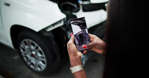 Person fotografiert einen Frontschaden an einem weißen Auto mit dem Smartphone.